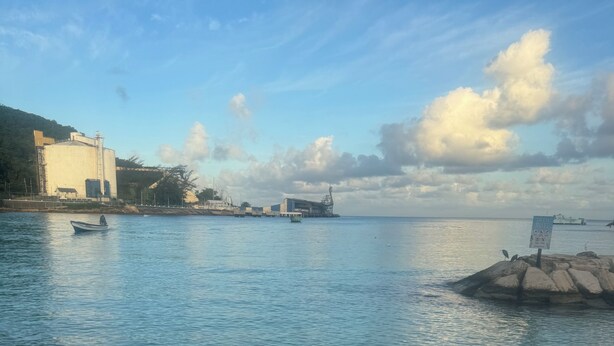 Una playa con aguas tranquilas, un bote y edificios industriales al fondo.