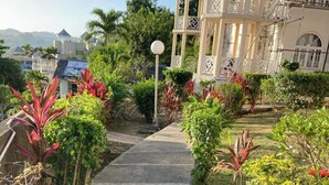 Un sendero conduce a un edificio de varios pisos con balcones, rodeado de vegetación exuberante y un cielo despejado.