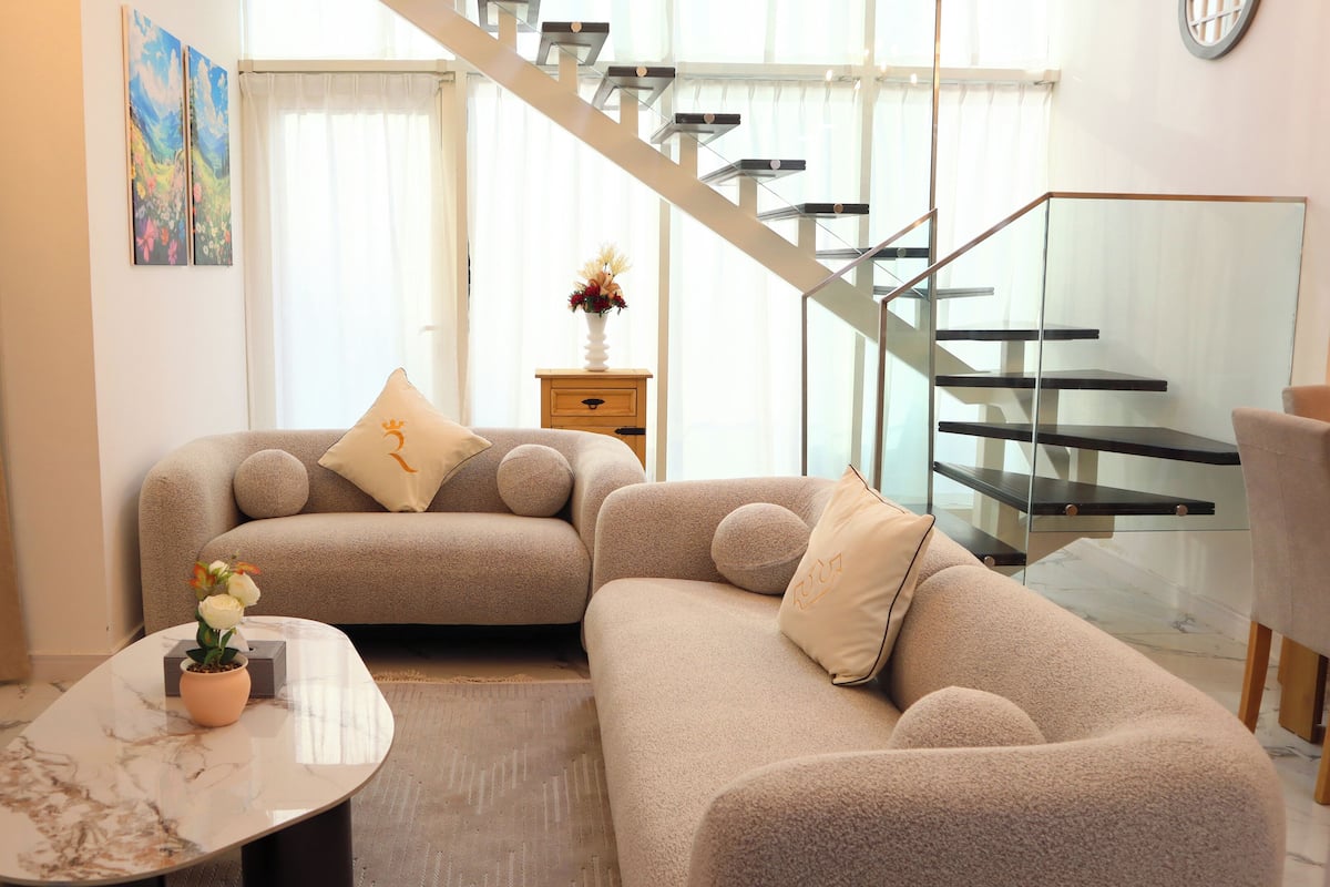 A modern living room with a sectional sofa, a marble coffee table, and a staircase with glass railings.