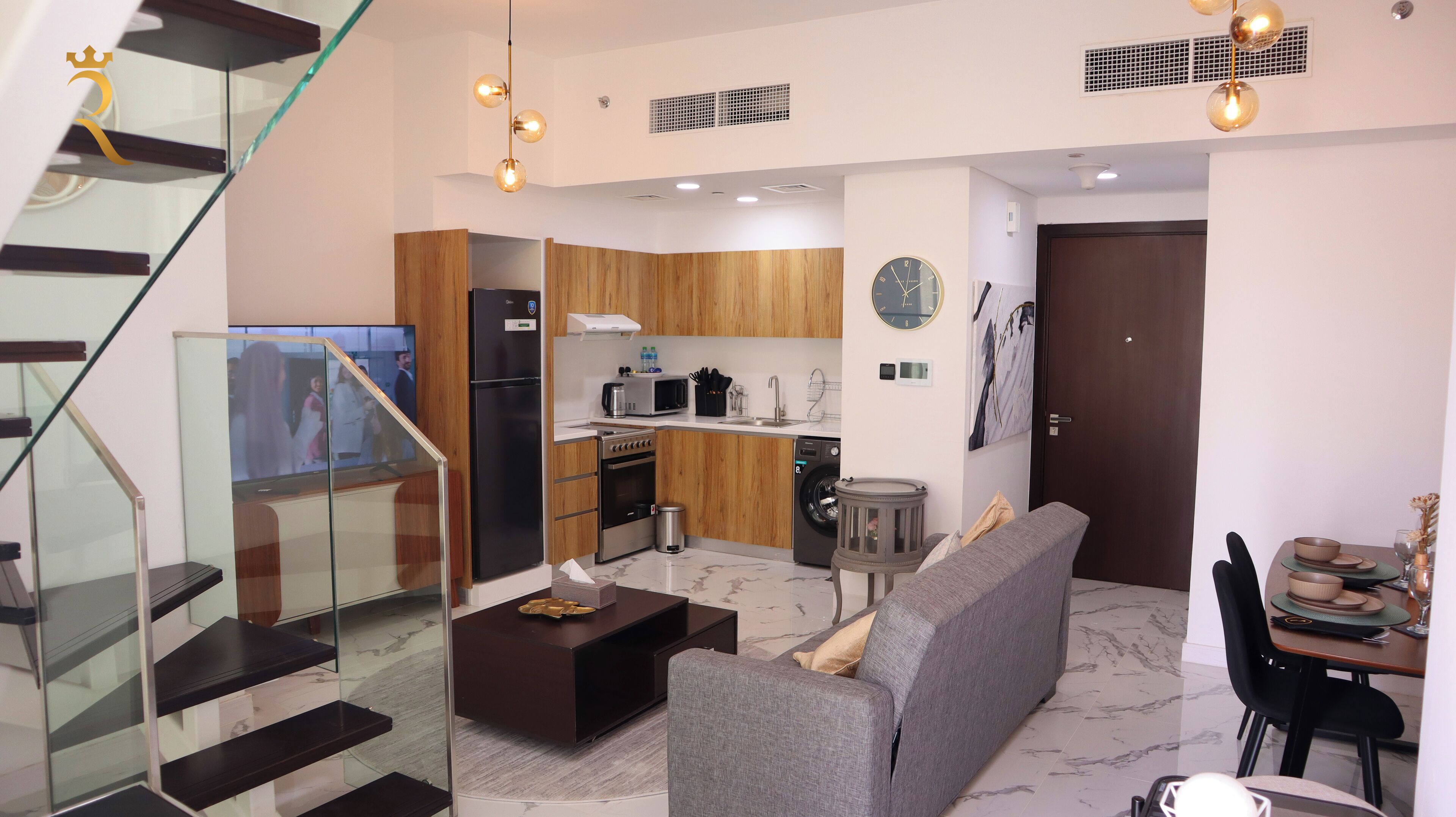 A modern kitchen with wooden cabinets, a built-in oven, and a microwave. There is a dining area with a gray sofa and a coffee table. A staircase with glass railings leads to an upper level.