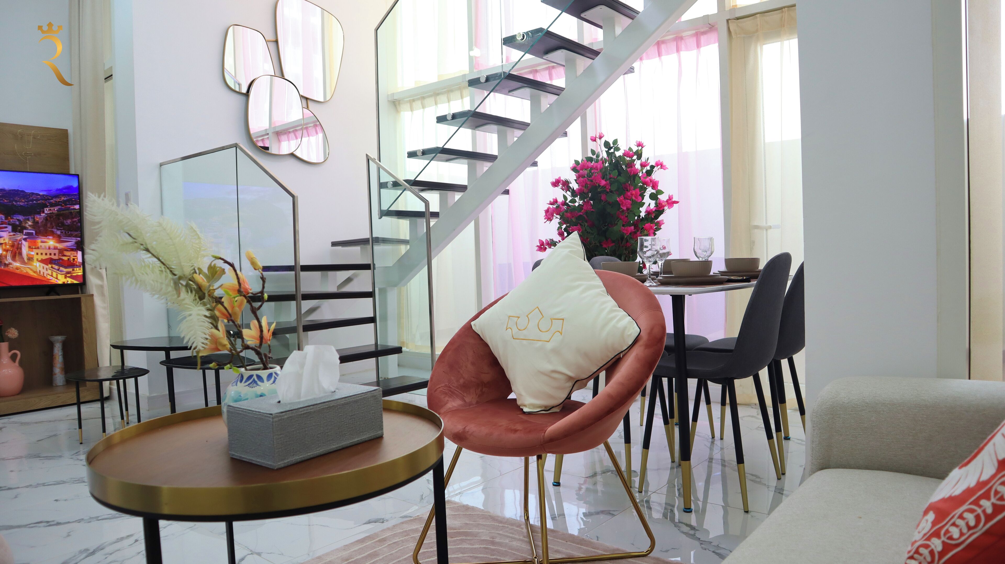 A modern living room with a sofa, a round coffee table, a pink chair, a dining area with a table and chairs, a staircase, and a TV on the left.