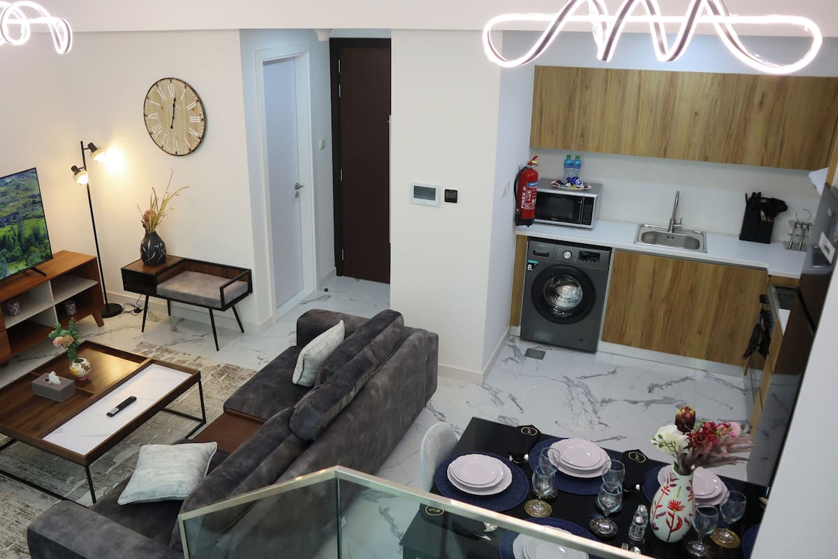 A modern living room with a sofa, coffee table, and TV. There's a kitchen with a washing machine and microwave.