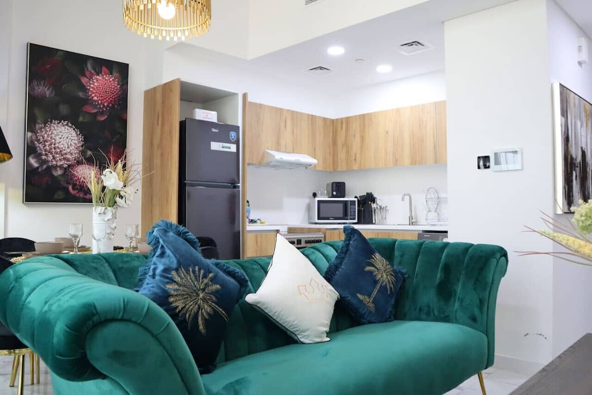 A modern living room with a teal sofa, a dining table, and a kitchen area with wooden cabinets and a microwave.