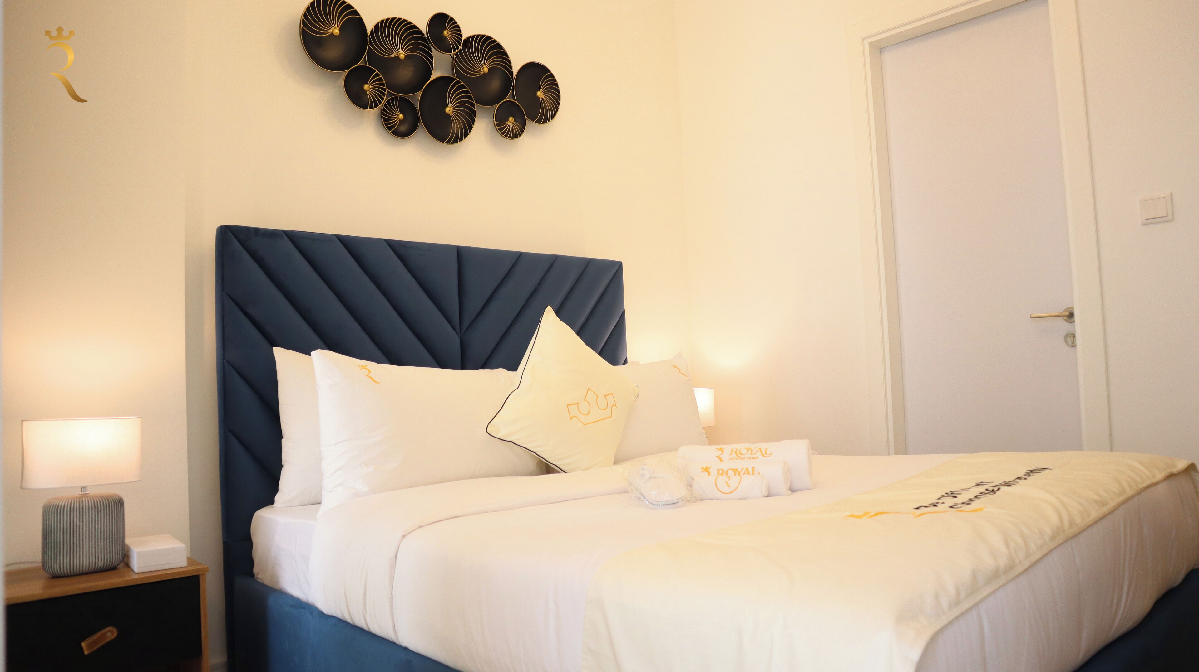 Een netjes opgemaakt bed met witte beddengoed en een donkerblauwe hoofdbord, een nachtkastje met een lamp, en een decoratie aan de muur erboven.