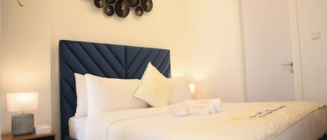 Een netjes opgemaakt bed met witte beddengoed en een donkerblauwe hoofdbord, een nachtkastje met een lamp, en een decoratie aan de muur erboven.