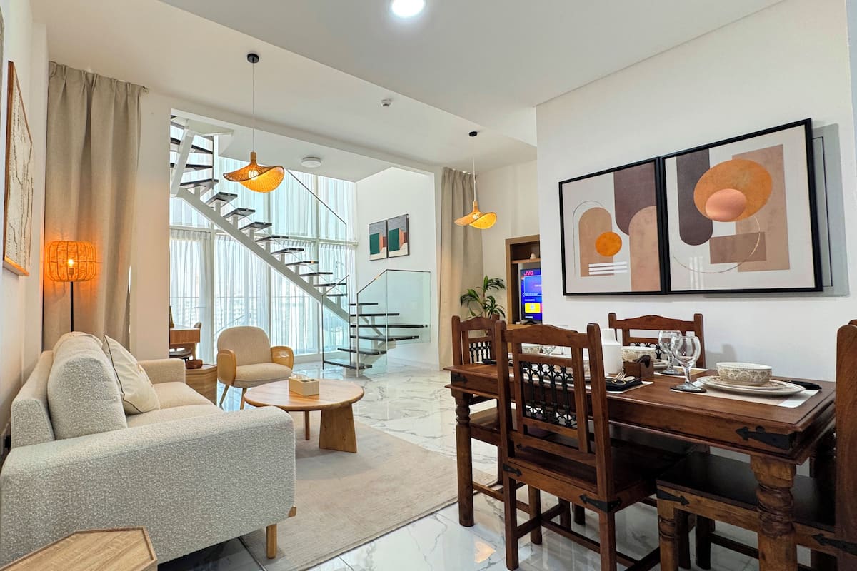 A modern living room with a sofa, dining table, chairs, and a staircase leading to an upper floor.
