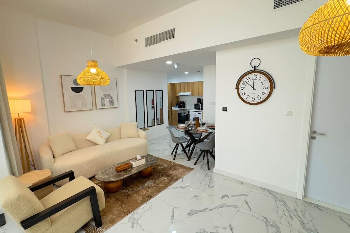 A modern living room with a sofa, coffee table, and chairs. There is a dining area with a table and chairs, and a kitchen in the background.