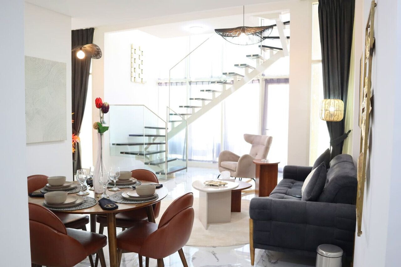 Una sala de estar moderna con un sofá gris oscuro, una mesa de centro y una zona de comedor con una escalera de cristal al fondo.