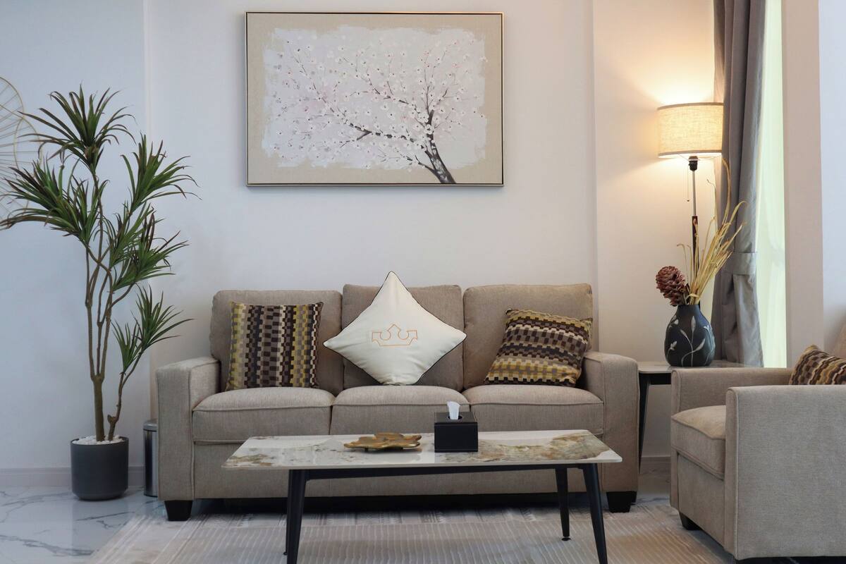 A living room with a beige sofa, two patterned pillows, a small table with a tissue box, a potted plant, and a framed artwork on the wall.