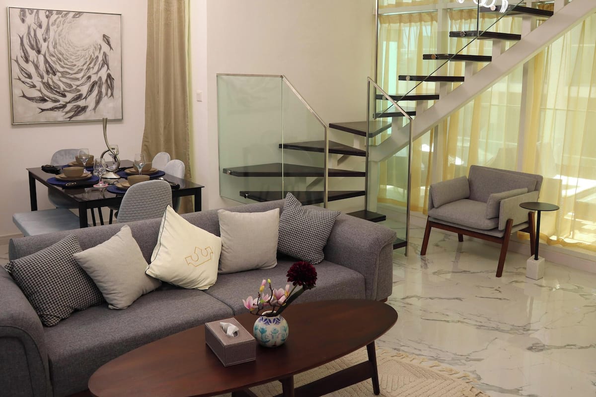 A modern living room with a grey sofa, a coffee table, and a vase with flowers. There is a dining area with a table and chairs, and a staircase with glass railings.