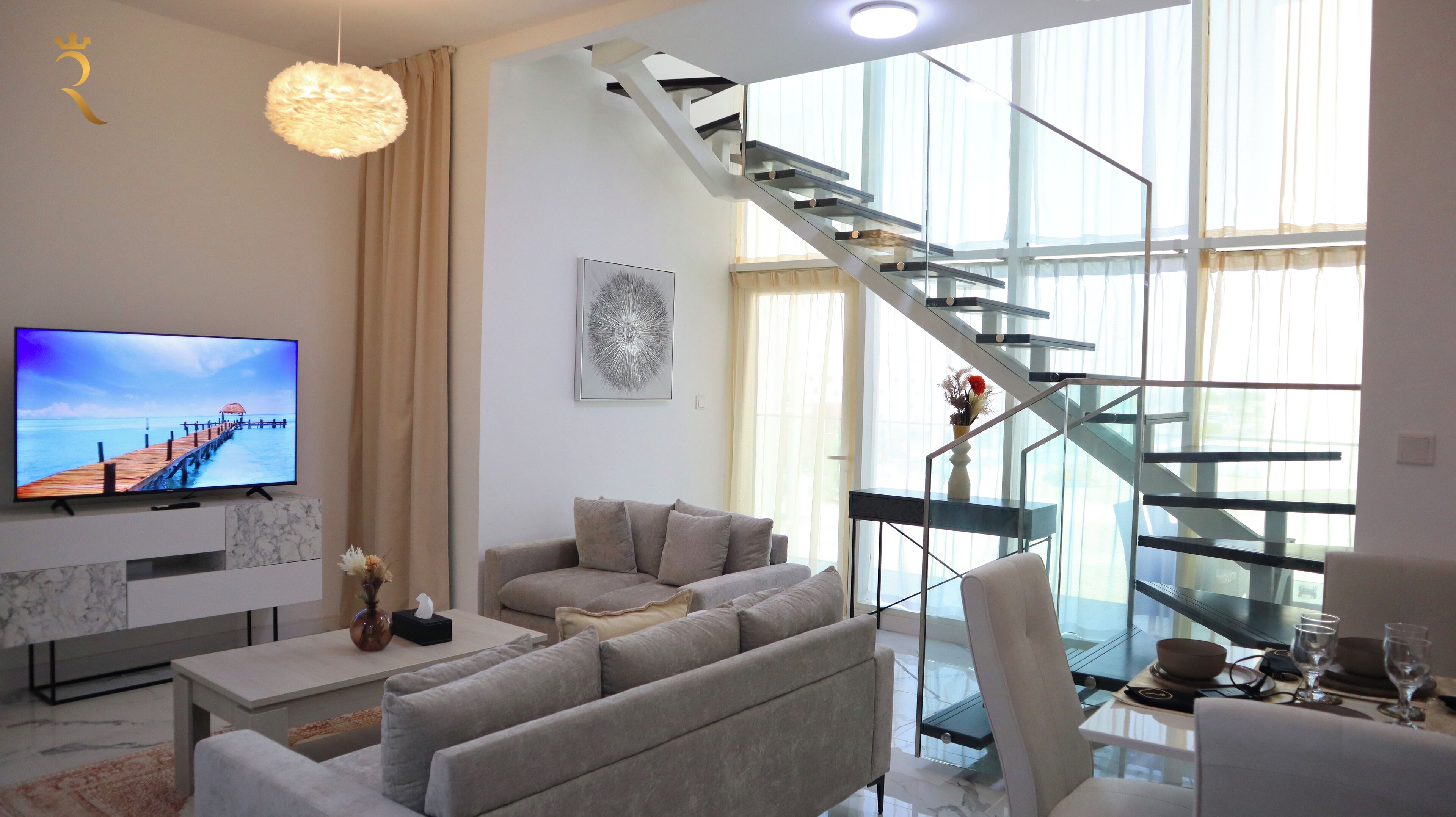 A modern living room with a large flat-screen TV, a sofa, a coffee table, and a glass staircase.