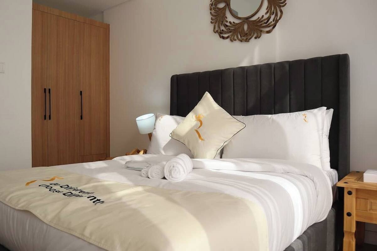 A neatly made bed with a white bedspread and a towel. There is a wooden wardrobe and a bedside table with a lamp.