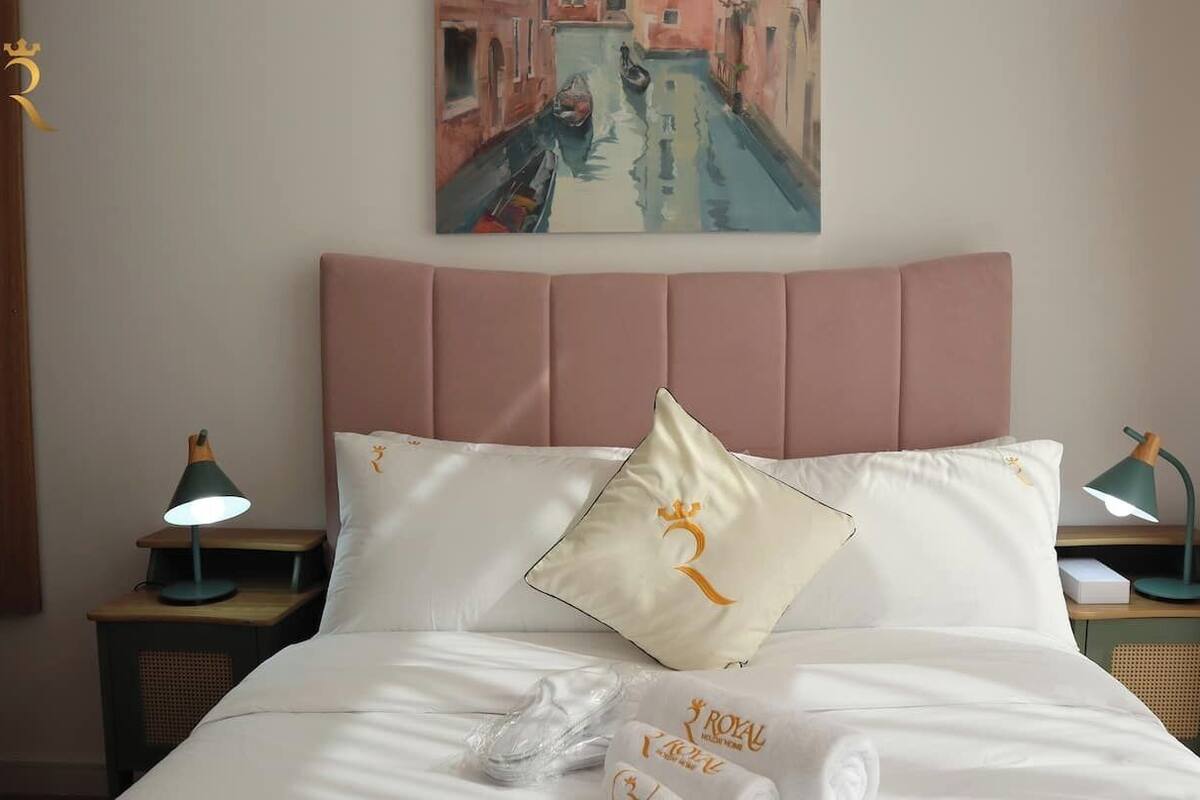 A hotel room with a bed, a painting of a canal, and a pillow with a crown symbol.