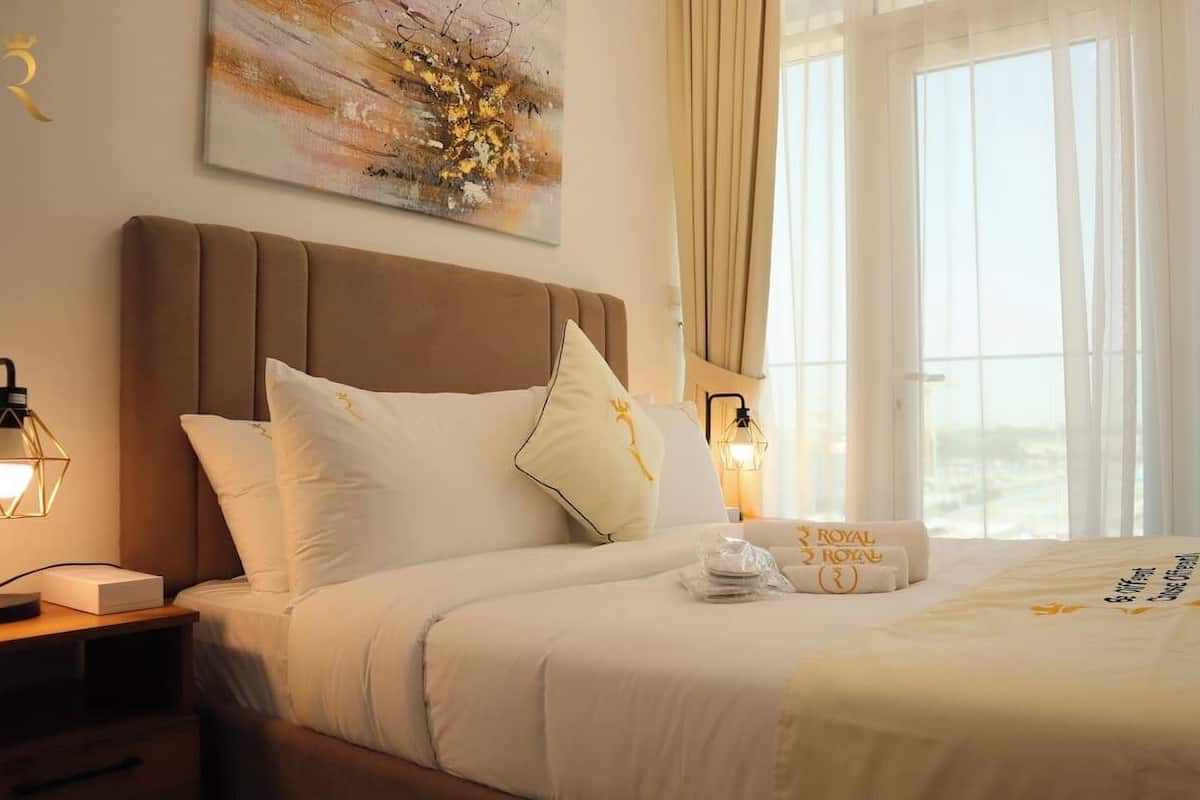 A hotel room with a large bed, two bedside lamps, a painting on the wall, and a view of the outdoors through the window.
