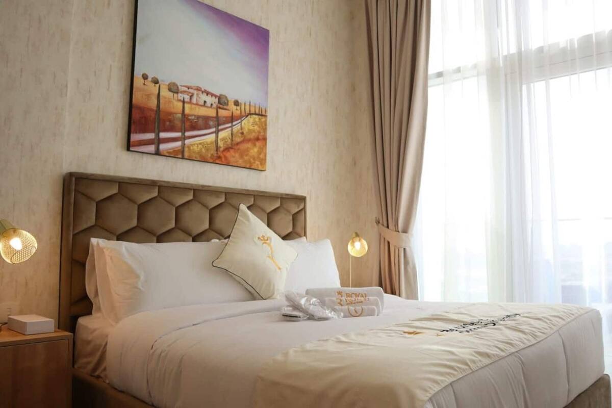 A neatly made bed with a geometric headboard, a painting above it, and a window with curtains.