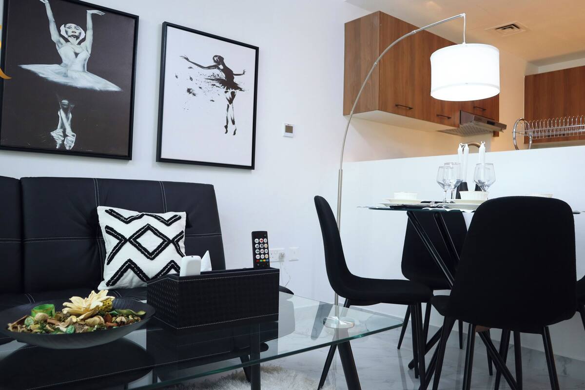 A modern living room with a black sofa, a glass coffee table, and a dining area with a white table and black chairs. There are two framed artworks on the wall, a remote control on the sofa, and a bowl of fruit on the table.