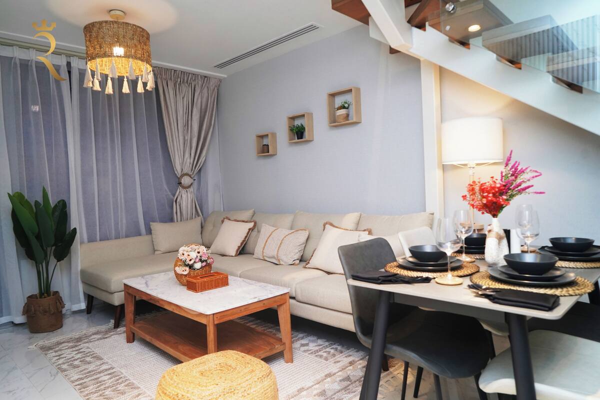 A modern living room with a sofa, coffee table, and dining area. There is a staircase in the background.