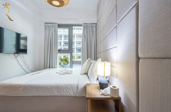 Habitación de hotel con una cama, un televisor de pantalla plana en la pared, una mesita de noche con lámpara y una ventana con cortinas.