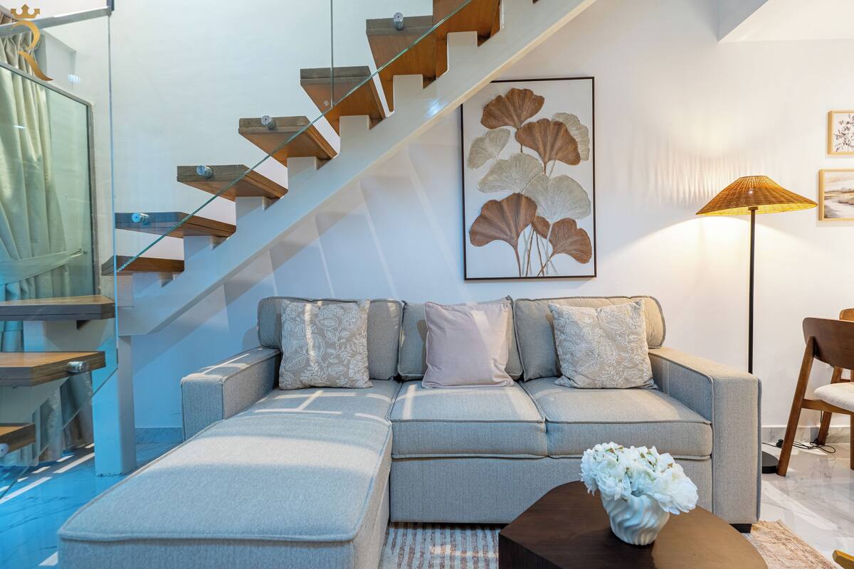 A modern living room with a grey sectional sofa, a glass staircase, and a framed botanical print on the wall.