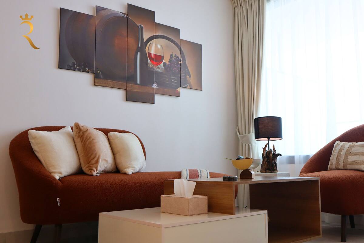 A modern living room with a brown sofa, a matching armchair, a small coffee table, and a lamp. There is a large window with curtains, and artwork on the wall.