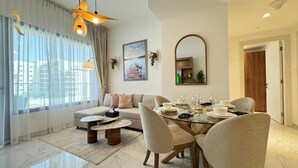 A modern living room with a glass dining table, beige sofa, and a large window with sheer curtains. - Masdar Oasis 1 Modern Apartmenst By Rhh (Abu Dhabi)