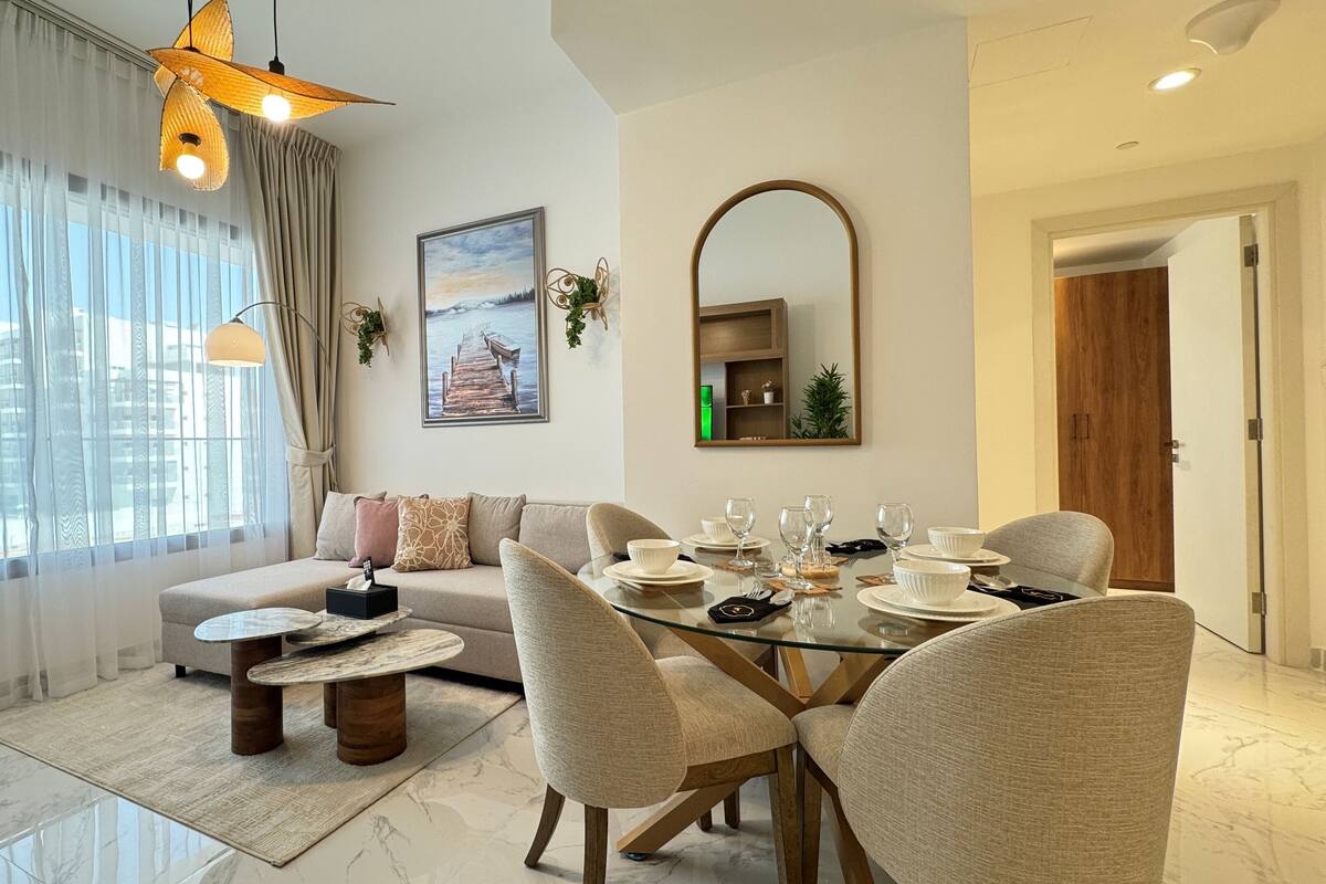 A modern living room with a glass dining table, beige sofa, and a large window with sheer curtains.