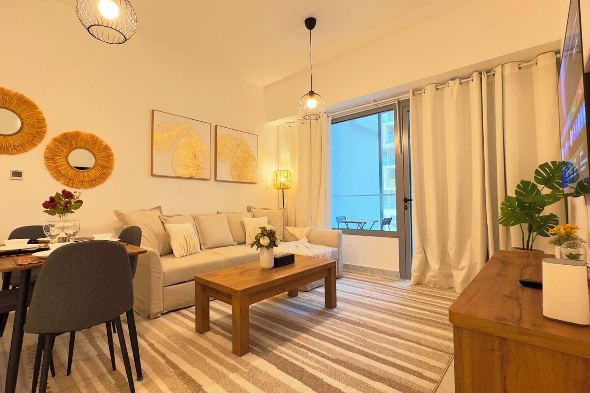 A modern living room with a sofa, coffee table, and dining area. There are two wall-mounted mirrors, a lamp, and a TV mounted on the wall.