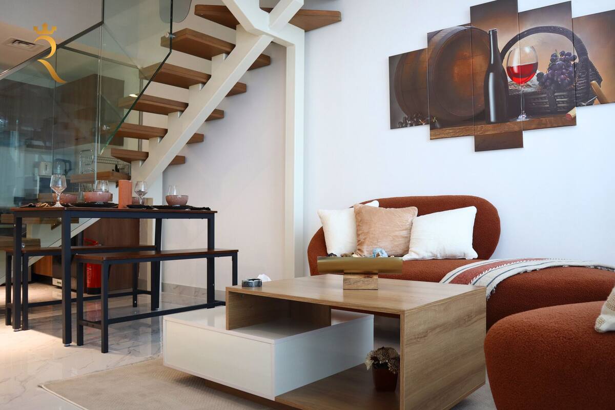 A modern living room with a staircase, a dining area with glass-top table, and a large wine-themed artwork on the wall.