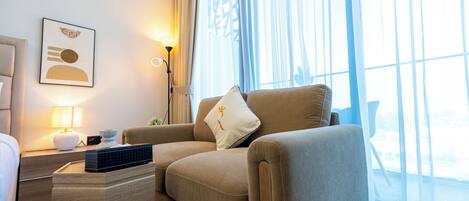 A modern hotel room with a beige sofa, a side table with a lamp, and a framed artwork on the wall.