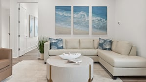 Un salon moderne avec un canapé d’angle beige, une table basse ronde et deux grandes peintures représentant des vagues océaniques, accrochées au mur.