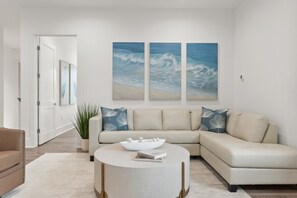 A modern living room with a beige sectional sofa, a round coffee table, and two large wall-mounted paintings of ocean waves. - 30A Turtle Cove Retreat Modern 8BR with Private Pool, Bikes, and Golf Cart (Santa Rosa Beach)