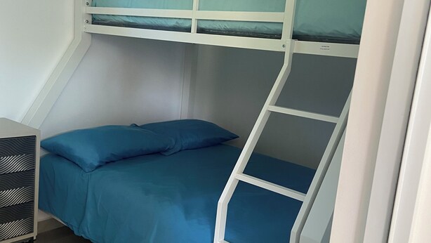 A bunk bed with a blue mattress and pillows, a white metal ladder, and a white wall with an air conditioning unit. - Coco and Playa camper (Aguada)