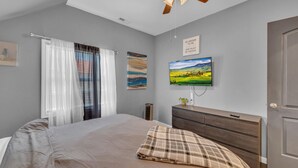 Une chambre à coucher avec un lit, un ventilateur de plafond, une télévision, une commode et une porte.