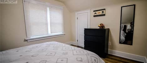 A bedroom with a bed, dresser, window with blinds, door, and a mirror on the wall.
