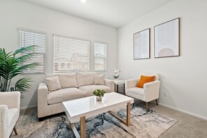 A living room with a beige sofa, a white coffee table, and a small side table with a plant. There are two windows with blinds, a potted plant, and two framed artworks on the wall.
