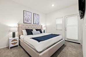 A bedroom with a tufted headboard, a bed with white bedding and a dark blue throw, two bedside tables with lamps, and two abstract wall art pieces.