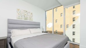Une chambre moderne avec un grand lit, une table de chevet et une fenêtre donnant sur un immeuble jaune.