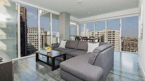 Un vaste salon doté d’un grand canapé d’angle gris, d’une table basse et offrant une vue sur la ville grâce à de grandes fenêtres du sol au plafond.
