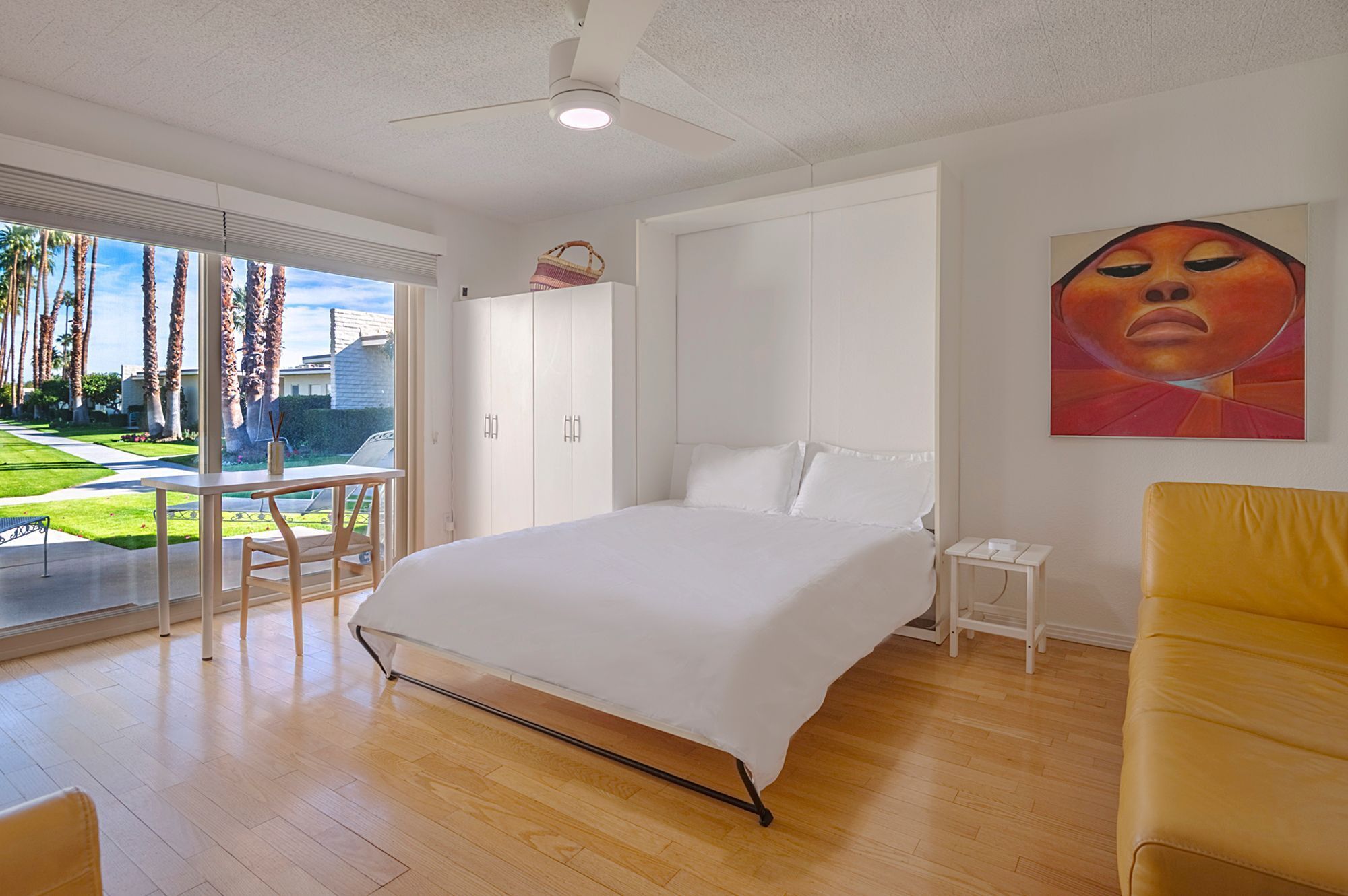 A bedroom with a bed, a desk, a chair, a small table, and a painting on the wall.