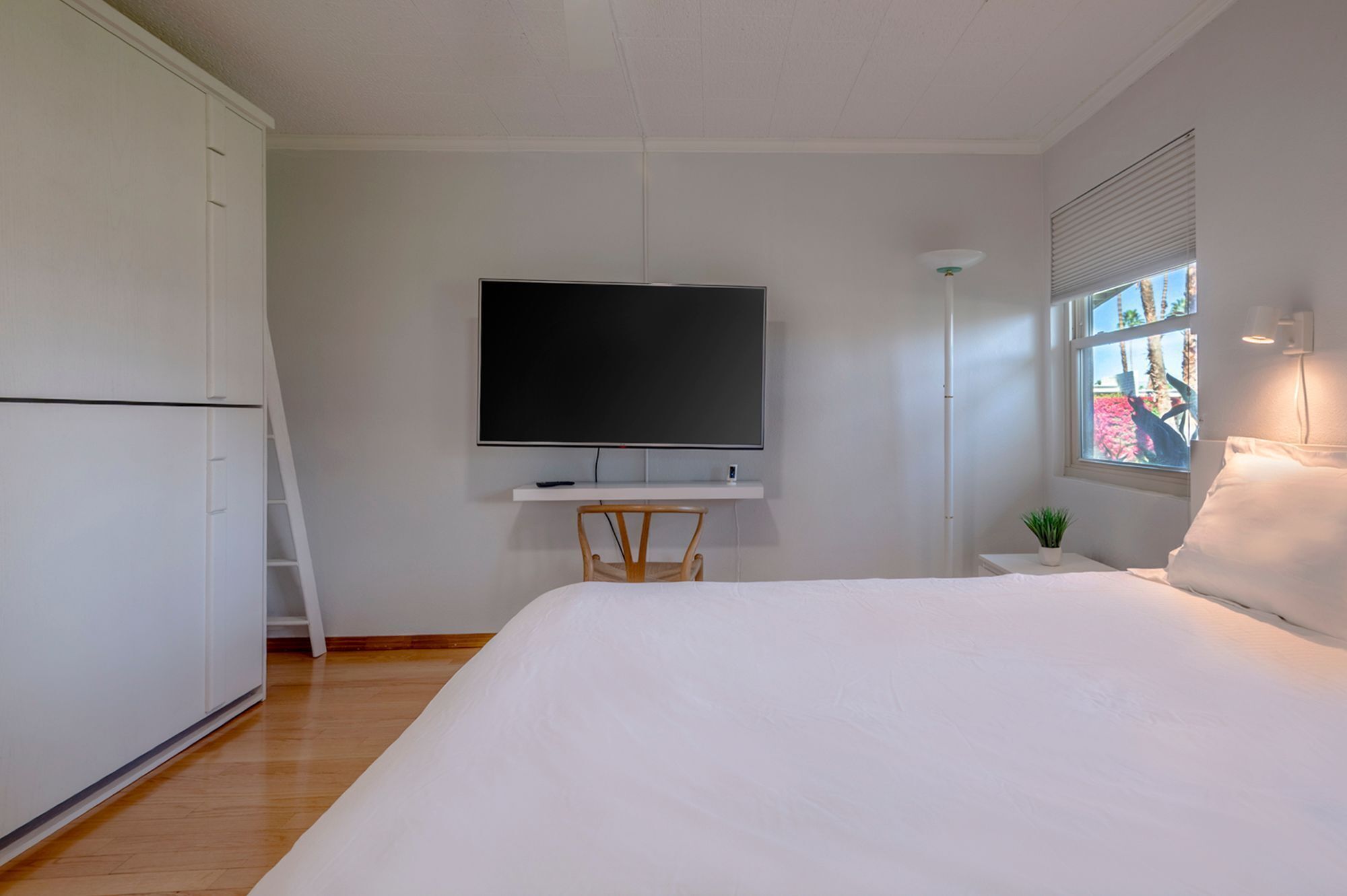 A bedroom with a bed, a TV mounted on the wall, a desk, a chair, a bedside table with a lamp, and a window with blinds.