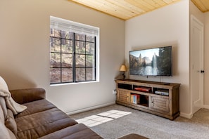 A living room with a large window, a flat-screen TV, a wooden entertainment unit, and a brown sofa. - Noble Estates Cabin, Luxe Forest Escape (Nutrioso City)