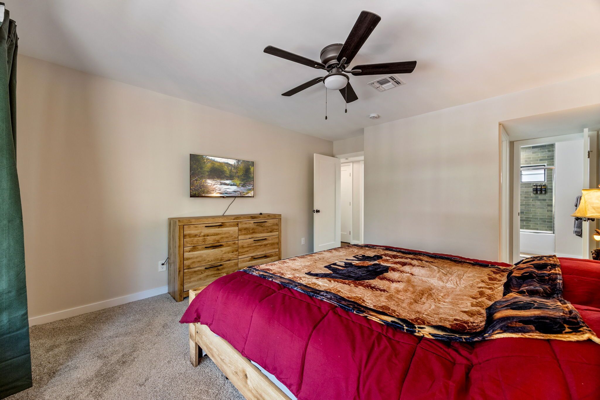 A bedroom with a bed, a ceiling fan, a wooden dresser, a painting on the wall, and a door leading to another room.