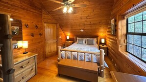 Een slaapkamer in een blokhut, met een bed, bedlampen, een houten dressoir en een raam met uitzicht op bomen.