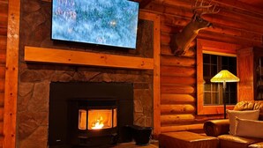 Een gezellig chaletinterieur met een stenen open haard, een opgehangen hertenhof, een televisie en een leren bank.