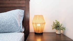 Ein Bett mit einem blumigen Kopfkissen, einer gewobenen Lampe und einem Blumentopf auf einem Nachttisch aus Holz.