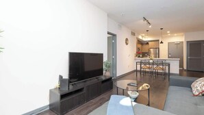 Ein modernes Wohnzimmer mit Flachbildfernseher, einer Couch, einem Couchtisch und einem Essbereich, im Hintergrund befindet sich eine Küche.