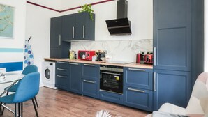 Eine moderne Küche mit dunkelblauen Schränken, einem Einbauofen und einer Mikrowelle. Eine Waschmaschine und ein Essbereich mit blauen Stühlen sind ebenfalls vorhanden.