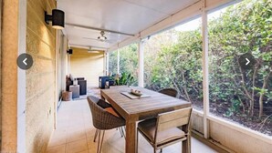 Une terrasse couverte avec une table et des chaises en bois, de grandes fenêtres et une vue sur la verdure.