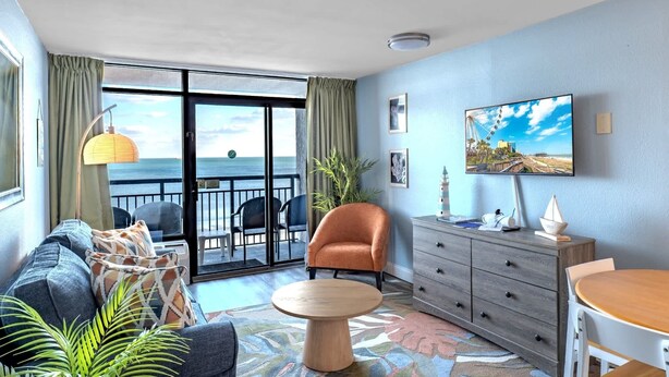 A room with a sofa, armchair, dining table, and a view of the sea through a sliding door. - Landmark 633 (Myrtle Beach)
