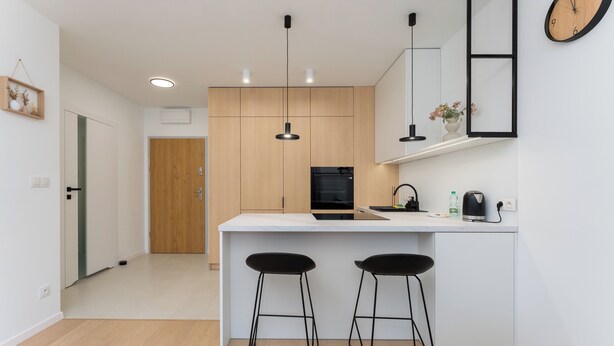 Een moderne keuken met een wit aanrecht, twee zwarte barkrukken, hanglampen en een zwarte koelkast.